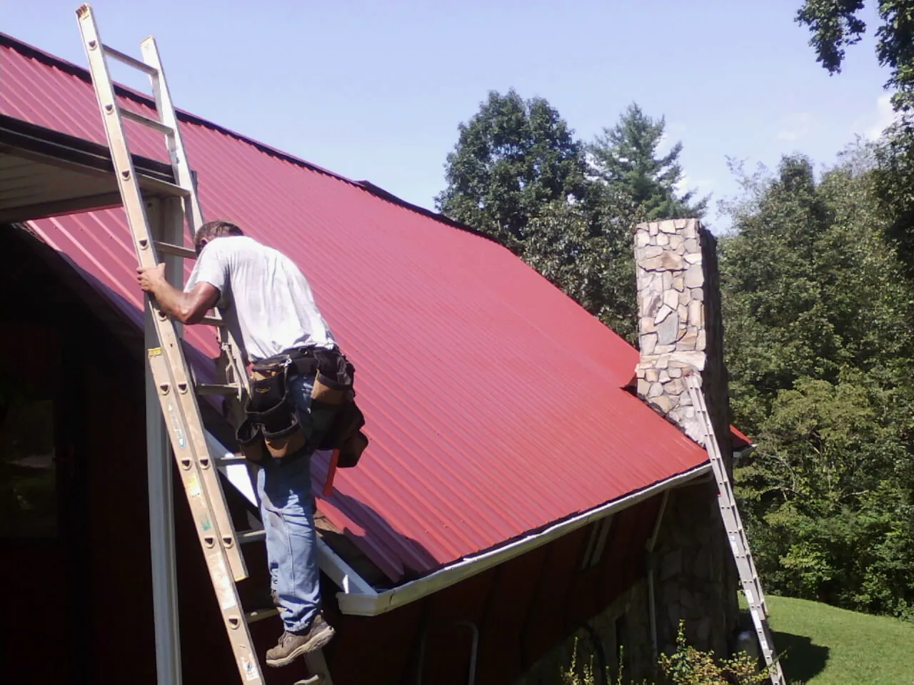 Roofer on a ladder beside a red metal roof and stone chimney — Soffit & Fascia Repair by Roofing Brothers in Fort Oglethorpe